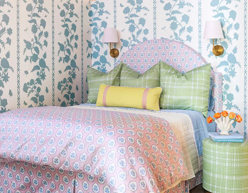 Pink and green bedding with matching upholstered headboard in room with blue wallpaper.